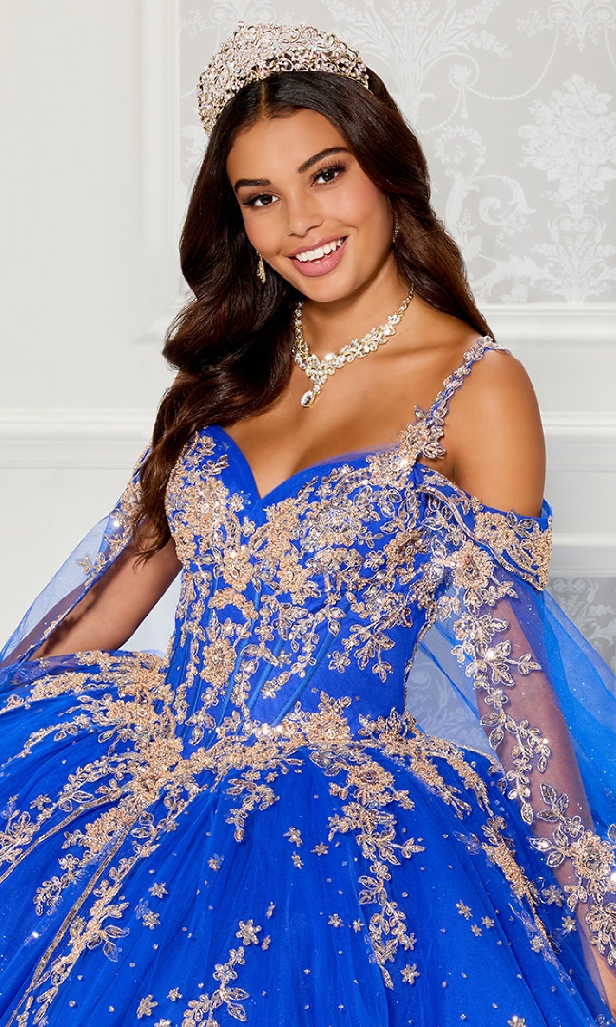 A young woman smiles in front of a light patterned background, wearing the Quinceanera Dress PR30087 By Princesa in royal blue with gold embroidery, a corset bodice, tiara, and necklace.